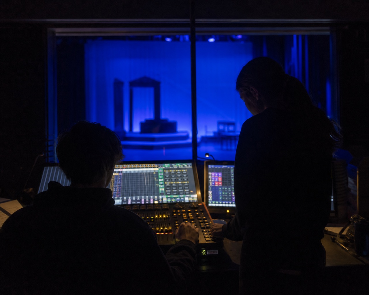 Two Vagabonds silhouetted at a lighting board, monitors and stage glowing through the booth window.